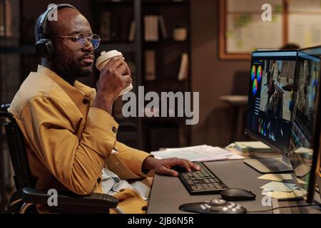 Colorista africano in cuffie con disabilità bere caffè durante la pausa al suo posto di lavoro in ufficio Foto Stock