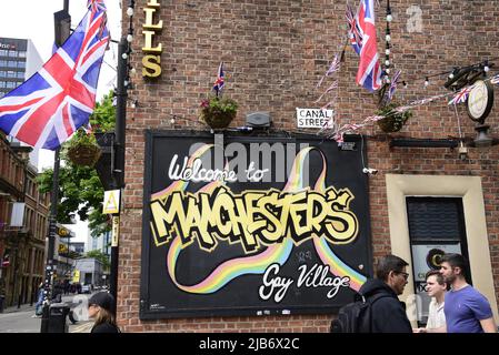 Manchester, Regno Unito, 3rd giugno 2022. La gente cammina oltre il cartello "Welcome to Manchester's Gay Village", Canal Street, Manchester, Regno Unito. Union Jack batte per celebrare il Giubileo. La vita quotidiana per le strade del centro di Manchester, Inghilterra, Regno Unito, Isole britanniche durante il Giubileo extra Bank Holiday Friday. Venerdì 3rd giugno è la festa ufficiale del Platinum Jubilee, una festa in più in occasione del 70th anniversario della Regina come monarca. Credit: Terry Waller/Alamy Live News Foto Stock