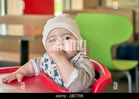 Una bambina con occhi marroni e grandi ciglia in un cappuccio leggero seduto su una sedia rossa per bambini che mangia una frutta. Foto Stock