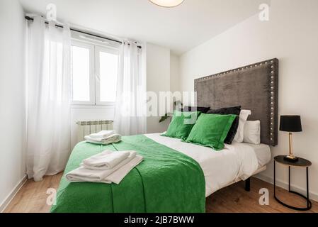 Camera da letto con un letto matrimoniale vestito in un impressionante copriletto verde e cuscini coordinati, testiera in tessuto imbottito e tende bianche sulla finestra Foto Stock
