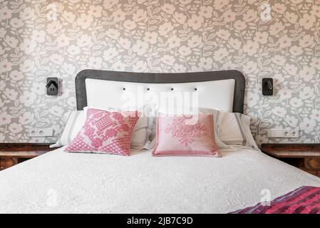 Testata della camera da letto rivestita in tessuto satinato grigio e bianco, cuscini rosa e parete con carta decorativa Foto Stock