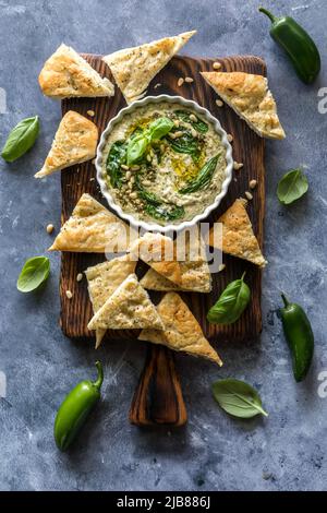 Pesto hummus con triangoli pita su un asse di legno, pronto per snacking. Foto Stock