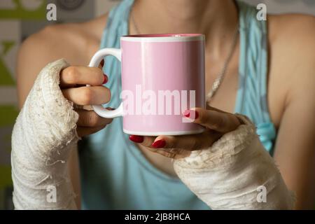 una ragazza con due braccia rotte in un cast tiene una tazza di tè in cucina per la prima colazione, braccia rotte in un cast, arti fratturate, lesioni Foto Stock
