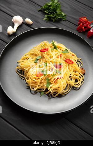 Pasta italiana fatta in casa cotta e in piedi su sfondo nero in un piatto nero accanto agli ingredienti aglio prezzemolo peperoncino. Scatto verticale Foto Stock