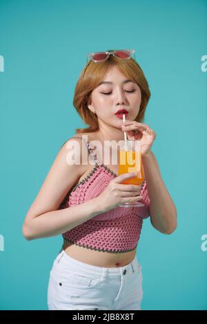 bella giovane ragazza bionda felice con top rosa e occhiali da sole colorati isolati su sfondo turchese tenendo succo d'arancia tazza bere e enj Foto Stock