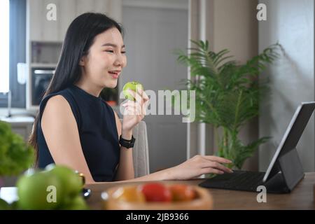 Ragazza allegra che mangia mela e usa un tablet in cucina mentre fa colazione. Foto Stock