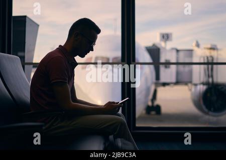 Silhouette di giovane uomo in attesa del volo. Il viaggiatore usa il telefono nel terminal dell'aeroporto. Foto Stock