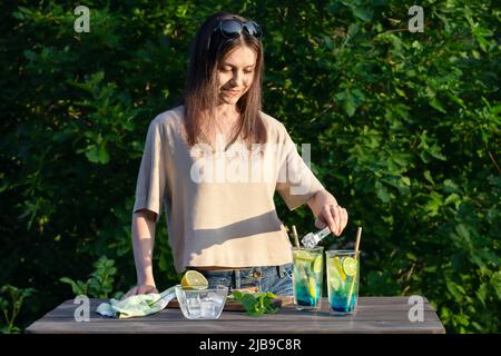 La ragazza fa i cocktail rinfrescanti estivi laguna blu. Processo di aggiunta di cubetti di ghiaccio ai bicchieri. Foto Stock