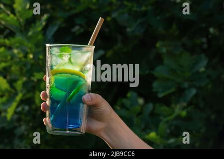 Bicchiere alto con un cocktail laguna blu in una mano femminile sullo sfondo del verde nel cortile. Messa a fuoco selettiva. Foto Stock