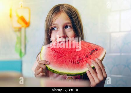 La bambina tiene una fetta del cocomero e la mangia. Un grande pezzo di cocomero rosso maturo nelle mani di una bambina su sfondo blu. Foto Stock