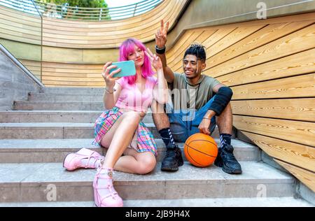 alternativa coppia diversificata appeso insieme seduto sulle scale della città facendo un selfie tenere smartphone. interracial adolescenti amici con palla Foto Stock