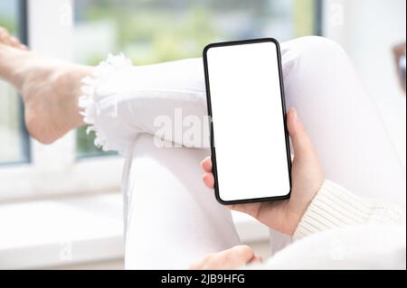 Donna in abiti bianchi si trova a gambe incrociate alla finestra a casa e utilizza il telefono cellulare o smartphone. USA l'app mobile shopping, controlla le notizie sui social media, textin Foto Stock