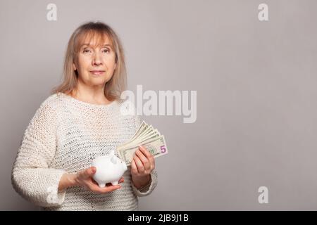 Felice sorridente donna anziana che ci tiene banconote in dollari e piggy banca su sfondo bianco. Foto Stock