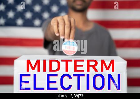 Il concetto di elezioni americane di Midterm del 2022 mostrando mettendo ho votato adesivo sulla scatola di voto con il segno di elezione di Midterm di fronte alla bandiera degli Stati Uniti Foto Stock