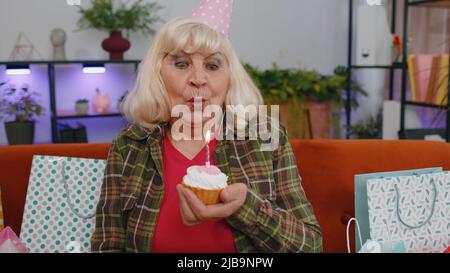Felice anziano anziano donna indossa festive compleanno cappello tenere cupcake fa desiderio gioioso congratularsi soffiando candela bruciante sulla torta. Nonna matura festeggia la festa sul divano con regali a casa Foto Stock