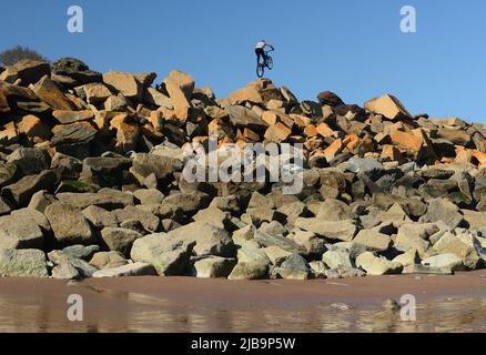 Un ciclista acrobatico che esegue acrobazie su armature di roccia multicolore, difesa del mare, visto dalla spiaggia sottostante con la bassa marea. Foto Stock