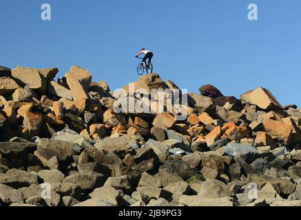 Un ciclista acrobatico che esegue acrobazie su armature di roccia multicolore, difesa del mare, visto dalla spiaggia sottostante con la bassa marea. Foto Stock