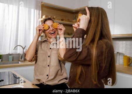 La mamma gioiosa e la figlia adulta si divertono in cucina durante il caffè, coprendo gli occhi con biscotti Foto Stock