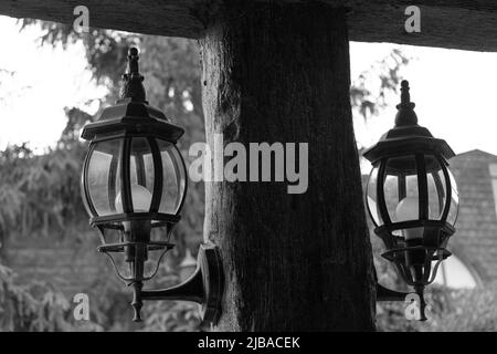 Lanterne metalliche di vecchio stile attaccate a colonne di legno, con alberi sullo sfondo, bianco e nero Foto Stock