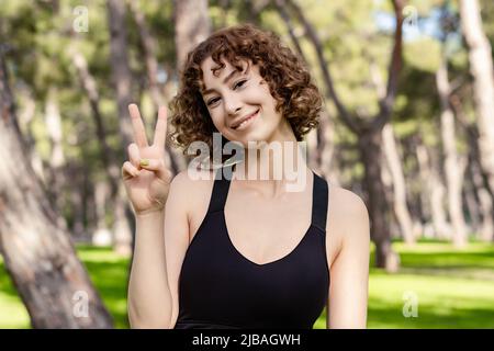 Ritratto di giovane donna rossa con reggiseno sportivo in piedi sul parco della città, all'aperto sorridendo con il volto felice che vana alla macchina fotografica facendo segno di vittoria o. Foto Stock