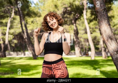 Bella donna rossa che indossa abiti sportivi in piedi sul parco cittadino, all'aperto felice ed eccitato facendo il gesto del vincitore con le braccia sollevate, sorridendo per s Foto Stock