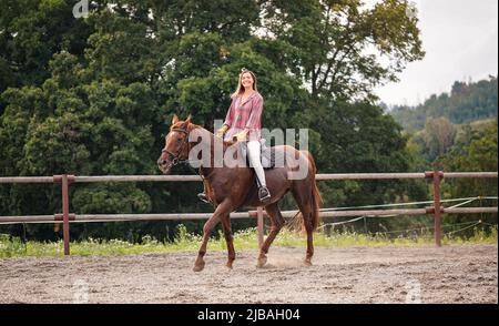 Giovane donna che indossa una camicia che va a cavallo marrone in paddock vicino a recinzione di legno, sfondo di alberi sfocati Foto Stock