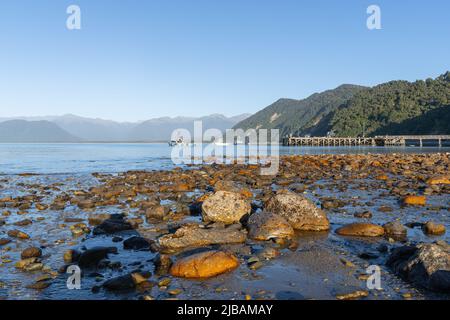 Luce del sole sul molo roccioso del litorale con le montagne circostanti mentre il sole tramonta attraverso Jackson Bay, South Island New Zealand. Foto Stock