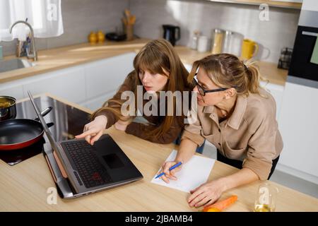 Madre e figlia sono alla ricerca di ricette di cottura seduti a un computer portatile in cucina Foto Stock