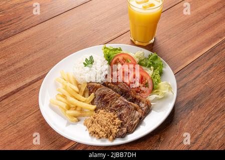 Bistecca di manzo con patatine fritte, fagioli e riso e un bicchiere di succo d'arancia su sfondo tavolo in legno, luce soffusa Foto Stock