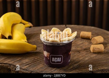 Deliziosa crema brasiliana Açaí, in una tazza di plastica con banana e arachidi macinate Topping, in un rustico sfondo di legno. Frullato estivo di acai Foto Stock