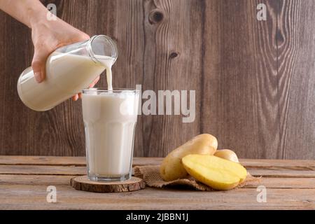 Il latte di patate vegane viene versato in un bicchiere e tuberi di patate su un rustico sfondo di legno. Prodotto di sostituzione del latte alternativo a base vegetale e privo di lattosio, Trend Foto Stock