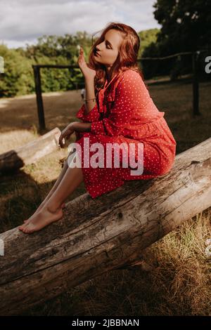 Bella giovane donna adulta in un abito rosso con puntini bianchi con capelli rossi seduti su un albero asciutto caduto e posando su una macchina fotografica. Modella femminile che la tocca Foto Stock