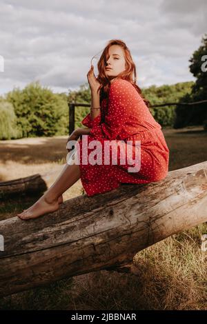 Bella giovane donna adulta in un abito rosso con puntini bianchi con capelli rossi seduti su un albero asciutto caduto e posando su una macchina fotografica. Modella femminile che la tocca Foto Stock