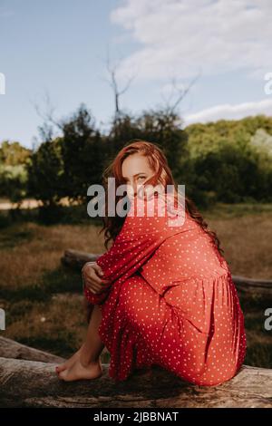 Bella giovane donna adulta in un abito rosso con puntini bianchi con capelli rossi seduti su un albero asciutto caduto e posando su una macchina fotografica. Modella femminile che la tocca Foto Stock