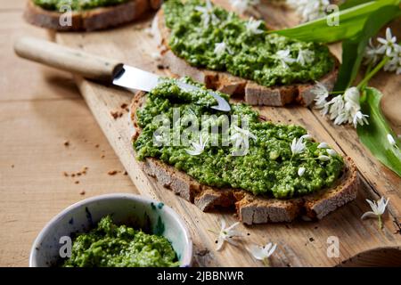 Pesto verde fresco con pane su sfondo ligneo con parmigiano, pinoli e pane, decorato con foglie e fiori di aglio selvatico. Foto Stock