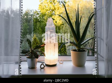 Ruvida grande selenite torre palo lampada illuminata in casa sul davanzale, foresta estiva sullo sfondo, spirituale casa arredamento accento e ambiente mo Foto Stock