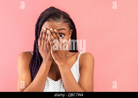 Ritratto di giovane donna nera terrorizzata in vestito coprendo il suo volto con le mani e sbirciando attraverso le dita su sfondo rosa Foto Stock