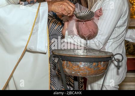 Un neonato viene battezzato nel rito religioso cattolico da un sacerdote vestito di bianco, versando acqua Santa sulla testa Foto Stock