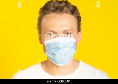 Bell'uomo biondo che indossa maschera medica prevenzione pandemica coronavirus o scimmietta. Bell'uomo in maglietta bianca e maschera medica su sfondo giallo. Panoramica, concetto di assistenza sanitaria. Foto Stock