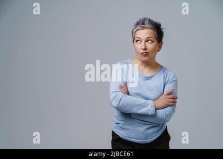 Donna matura, pensosa, in dubbio, con capelli grigi nel 50s in posa con le mani piegate e spazio copia a sinistra isolato su sfondo bianco. Luogo per il posizionamento del prodotto. Bellezza invecchiata. Immagine a toni. Foto Stock