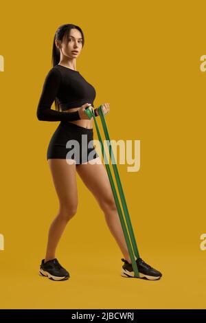 Bella ragazza sportiva in abbigliamento sportivo nero che si esercita con elastico resistente in studio. Vista laterale dell'atleta femminile che guarda la fotocamera durante l'allenamento, isolata su sfondo arancione. Concetto di fitness. Foto Stock