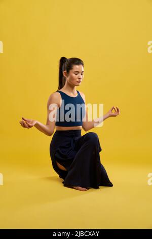 Calmo donna che fa squatting equilibrio posa con le mani mudra, esercitando yoga indoor. Vista frontale della giovane ragazza in nero top meditating, isolato su sfondo giallo, spazio copia. Concetto di meditazione. Foto Stock