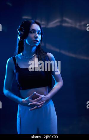 Donna sognante che indossa cuffie nere in piedi, stringendo le mani all'interno. Vista frontale della tranquilla femmina dj in cima al raccolto ascolto di musica, illuminata da luce blu scuro in studio. Concetto di gesturing. Foto Stock
