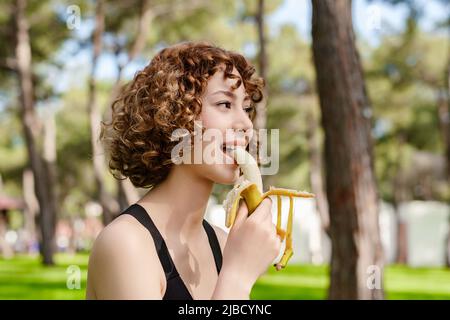 Bella donna rossa che indossa abiti sportivi in piedi sul parco della città, mangiare all'aperto banana sana per forza ed energia. Sport all'aperto e guarire Foto Stock