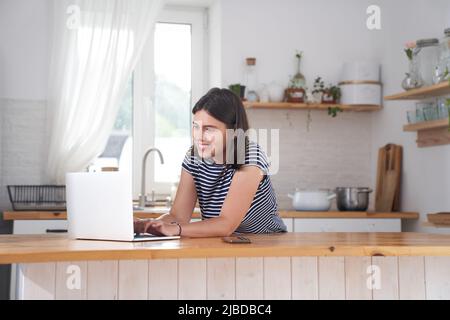 Una donna in cucina al computer con un sorriso. Una donna digita su un computer e lavora da casa o comunica sui social network. Foto di alta qualità Foto Stock