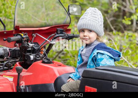 Un felice ragazzo di tre anni è visto nel sedile del conducente di un veicolo rosso per tutti i terreni con manubrio in campagna. Con sfondo di fogliame sfocato. Foto Stock