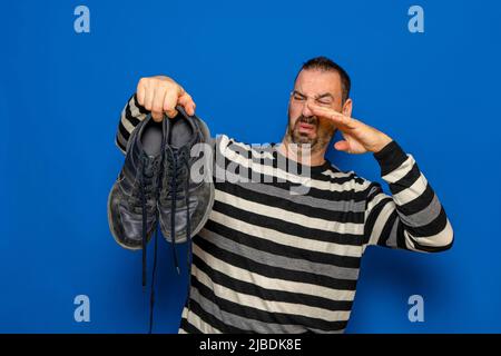 Un uomo tiene le sneakers nelle sue mani. Odore sgradevole. Gambe stinky, sudorazione eccessiva dei piedi. Isolato su sfondo blu studio. Foto Stock