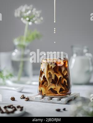 Caffè ghiacciato in un bicchiere trasparente con ghiaccio e latte su sfondo grigio. La caduta del latte si trasforma in un bicchiere di caffè. Foto Stock