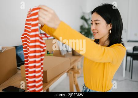 Sorridente giovane donna asiatica che disimballa i vestiti mentre si siede alla scrivania Foto Stock