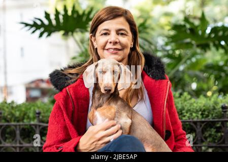 Donna latina in una giacca rossa che tiene il suo cane e guarda la macchina fotografica Foto Stock